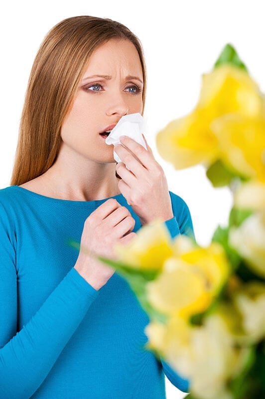 woman with a tissue in front of her face in a blue shirt with flowers in the foreground, representing allergies