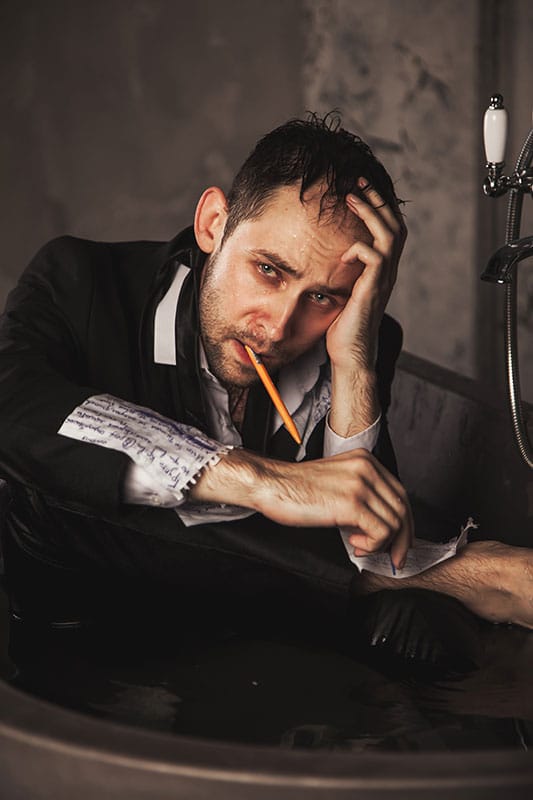 Frazzled looking man with a pen in his mouth, paper with writing on his arm, writing on a piece of paper on his leg, representing depression